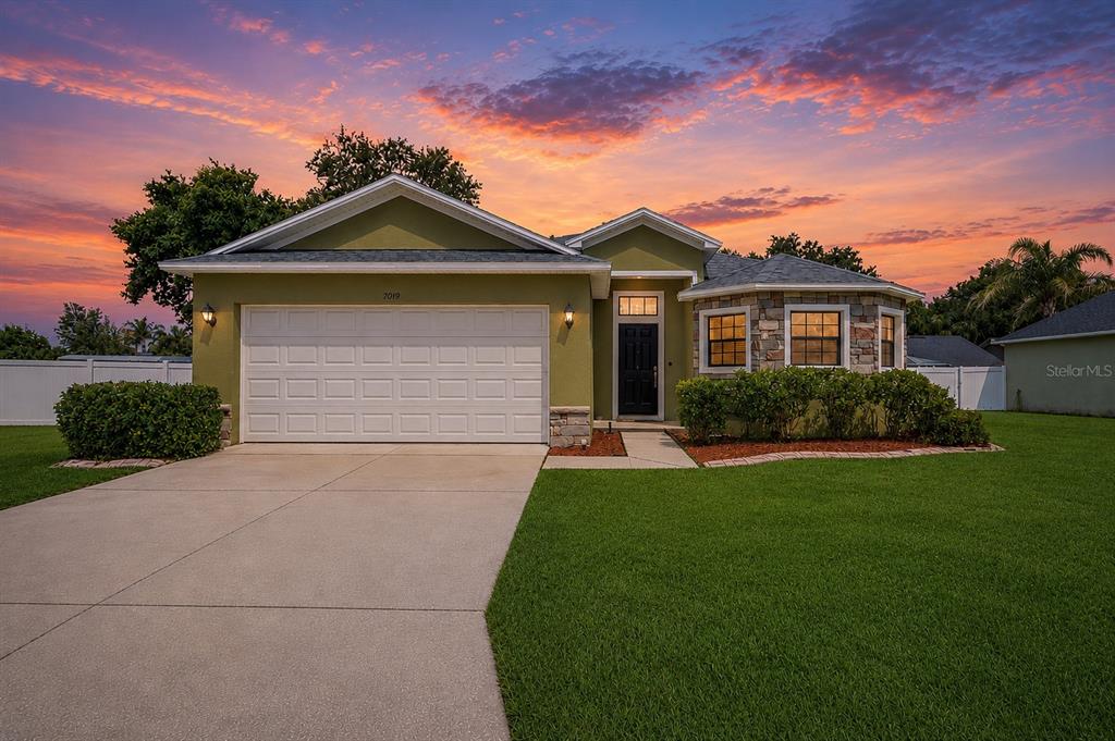 A single-story house with a green exterior, stone accents, and a two-car garage at sunset — 7019 HEATHERBROOK DRIVE, LAKELAND, FL