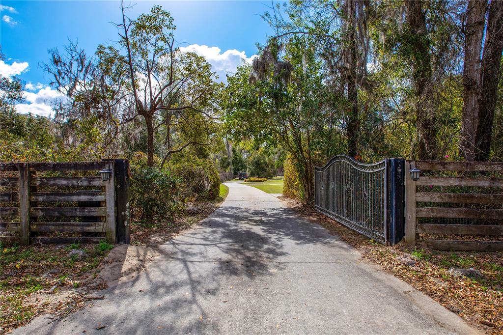 A paved driveway leads through an open metal gate flanked by wooden fences and trees — 12182 SW STATE ROAD 45, ARCHER, FL