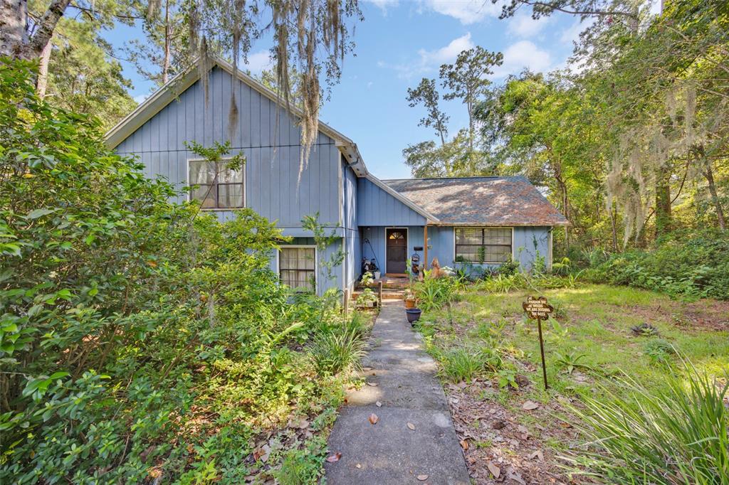 A two-story blue house with vertical siding stands behind a concrete walkway and overgrown yard — 7607 NW 42ND AVENUE, GAINESVILLE, FL