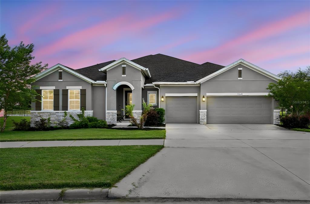 A single-story gray house with a three-car garage and stone accents under a sunset sky — 17060 SECRET HOLLOW LOOP, CLERMONT, FL