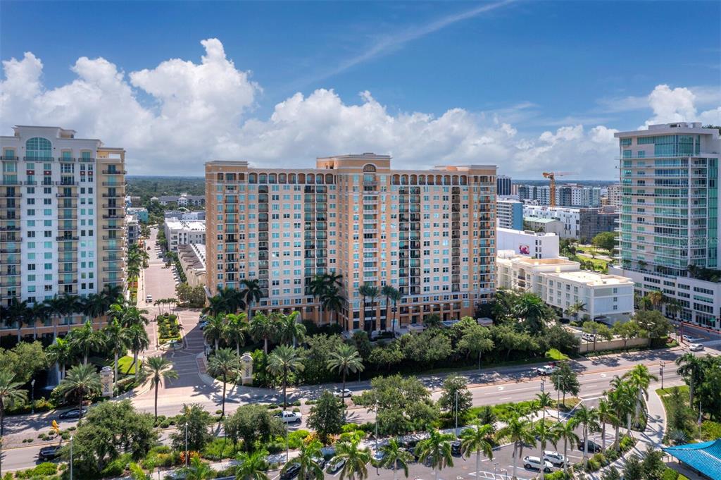 An aerial view shows a large, tan multi-story apartment building surrounded by palm trees and roads — 750 N TAMIAMI TRAIL UNIT 906, SARASOTA, FL