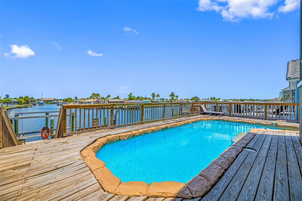 An outdoor swimming pool with a rock border sits on a wooden deck overlooking water — 861 SYMPHONY ISLES BOULEVARD, APOLLO BEACH, FL