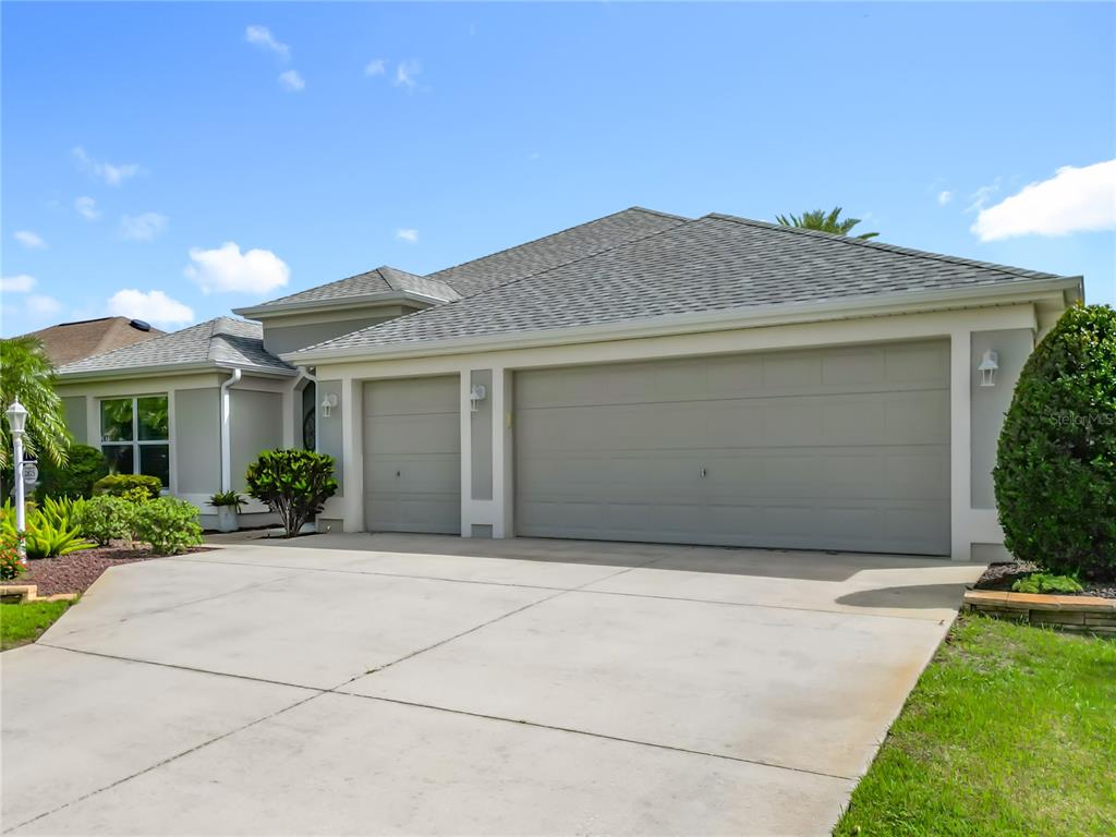A light-colored house exterior features a three-car garage and a concrete driveway — 2875 RAIN LILY LOOP, THE VILLAGES, FL