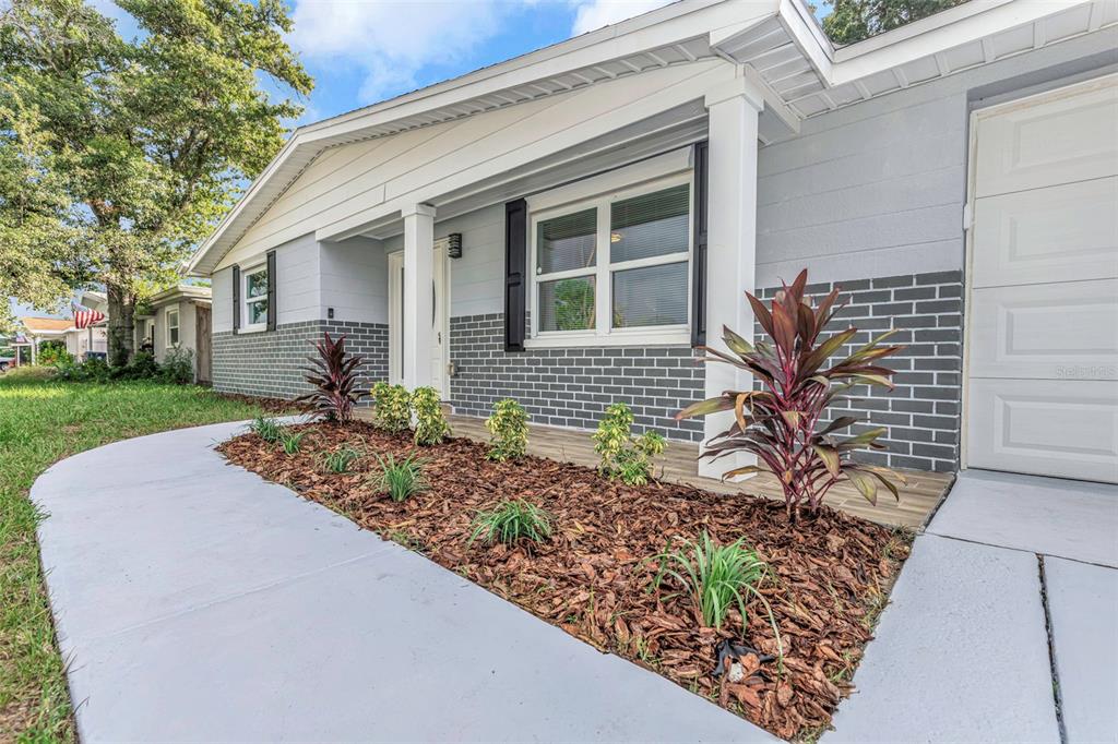 A gray house exterior features a brick facade, a covered porch, and a landscaped garden bed — 2332 CHANCERY DRIVE, HOLIDAY, FL