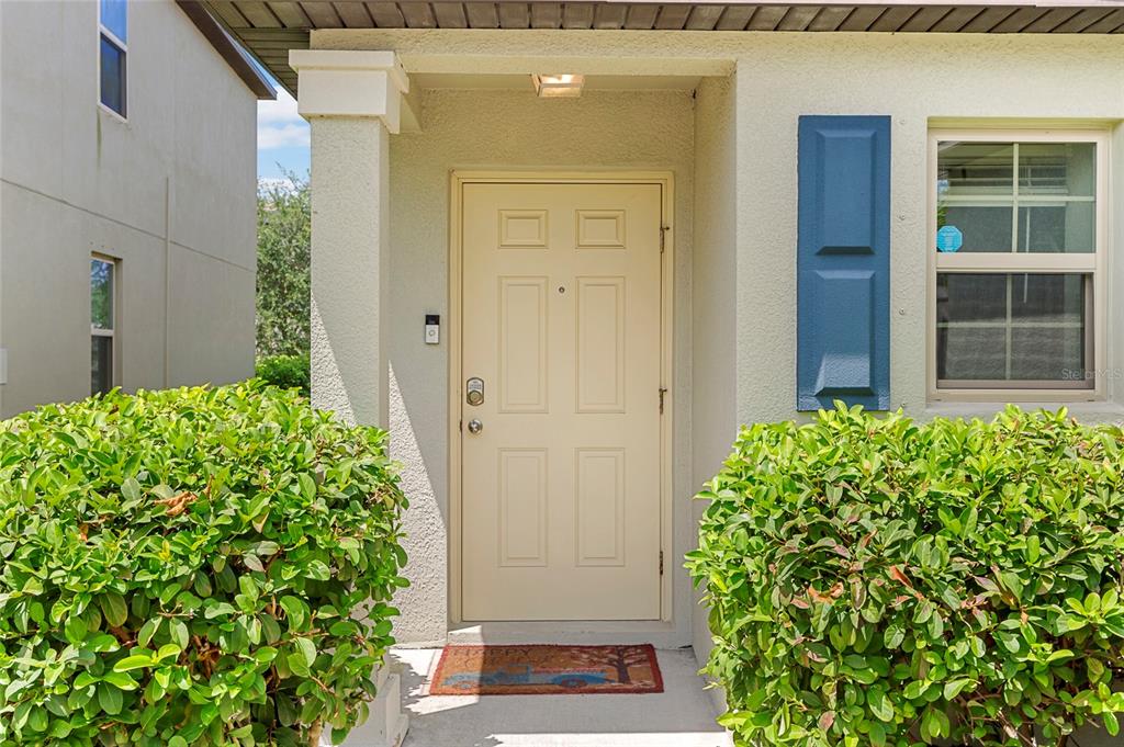 A beige front door with a doorbell camera is flanked by two large green bushes — 1719 FRED IVES STREET, RUSKIN, FL