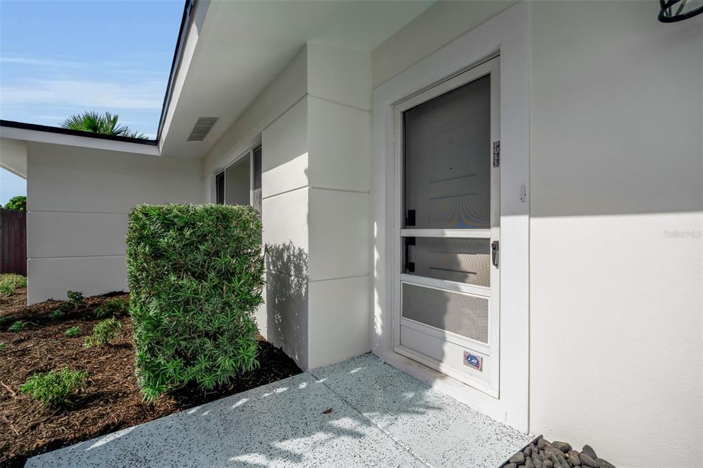 A white exterior wall features a storm door leading to a covered entryway with landscaping — 2769 WISTERIA PLACE, SARASOTA, FL