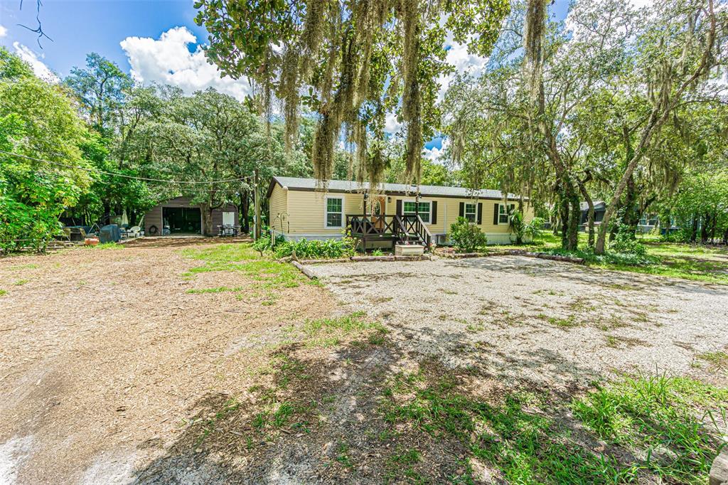 A single-story yellow mobile home sits on a gravel lot surrounded by large oak trees — 11420 ENGLISH WALNUT STREET, HUDSON, FL