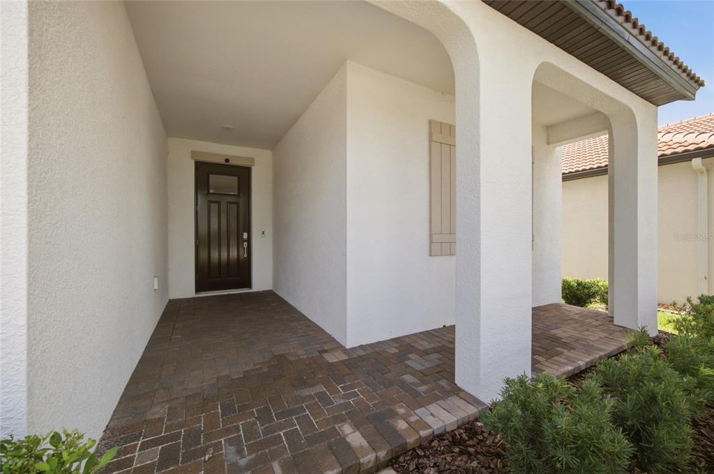 A covered entryway with a dark front door, white stucco walls, and brick paver flooring — 17218 STAR BANKS STREET, WIMAUMA, FL