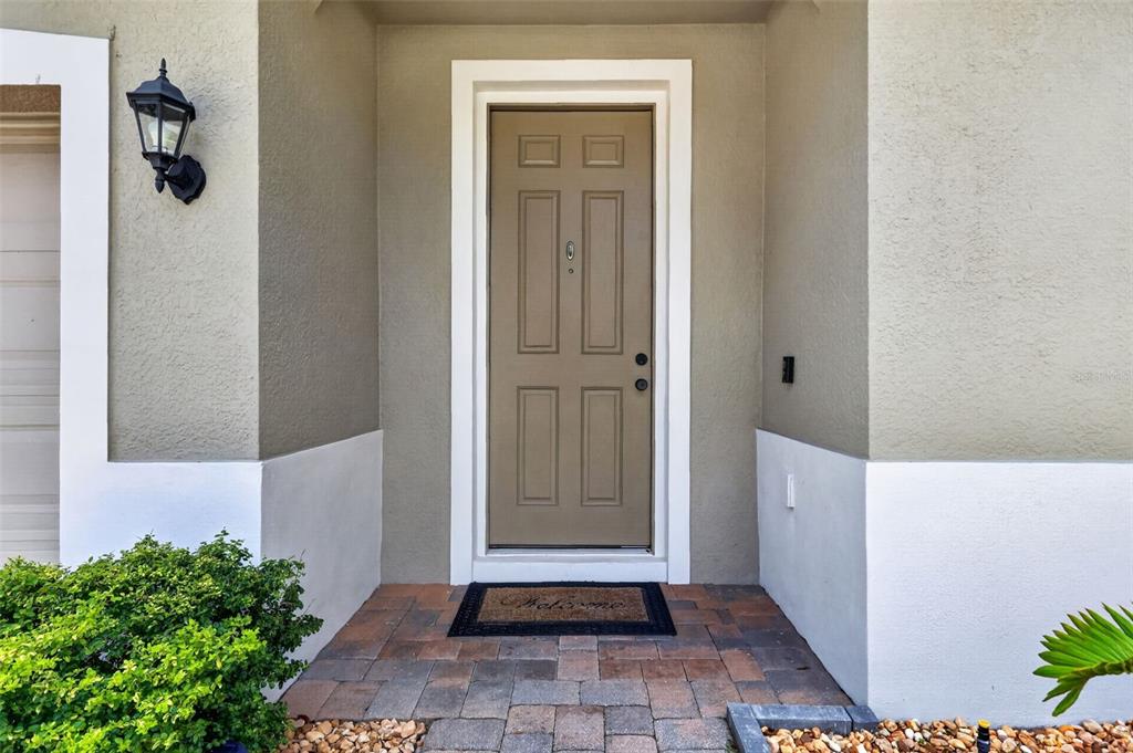 A tan front door with a welcome mat sits under a porch with stone pavers — 1450 OSPREY RIDGE DRIVE, EUSTIS, FL