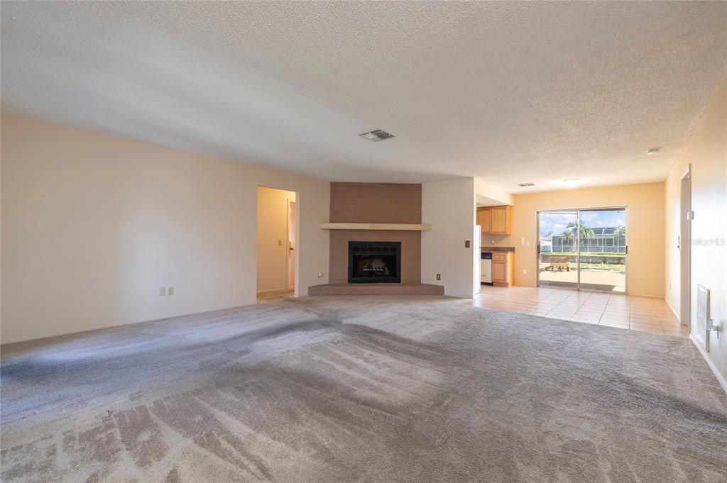 A living room with beige carpet, a corner brick fireplace, and an adjacent tiled kitchen area — 6355 102ND TERRACE N, PINELLAS PARK, FL