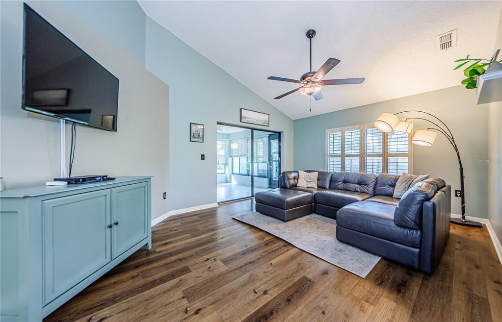 A living room features a dark leather sectional, wood-look flooring, and a wall-mounted television — 3647 MUIRFIELD COURT, NEW PORT RICHEY, FL