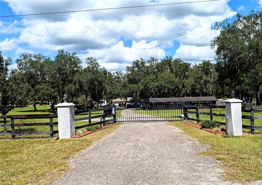 A gravel driveway leads to a gated entrance with stone pillars and wooden fencing — 10871 SE COUNTY ROAD 326, MORRISTON, FL