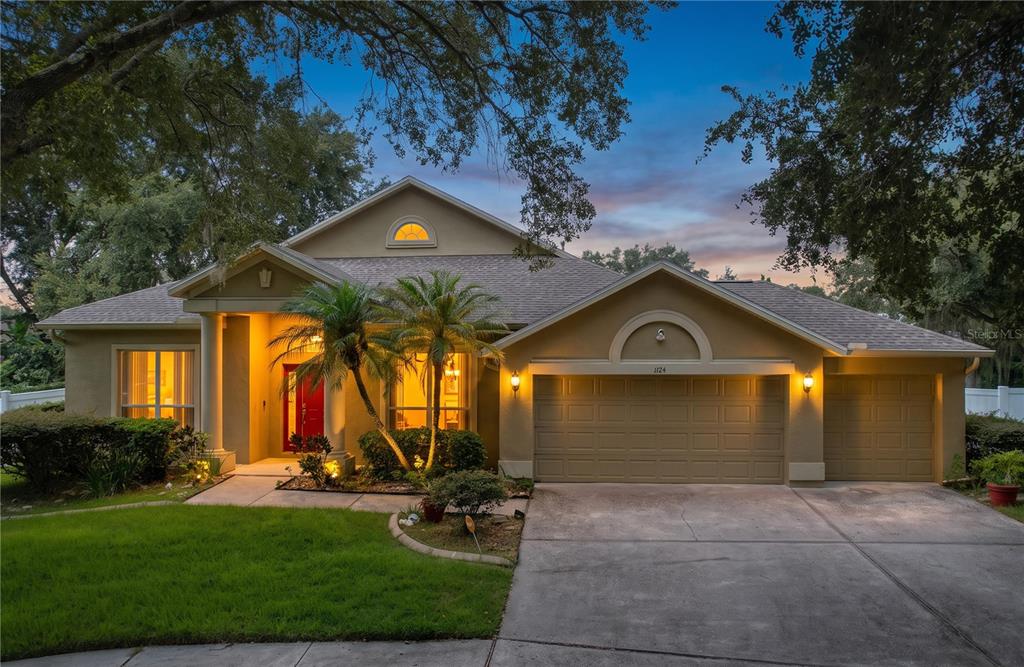 A single-story house with a tan stucco exterior, a red front door, and a three-car garage — 1124 SPLIT SILK STREET, VALRICO, FL