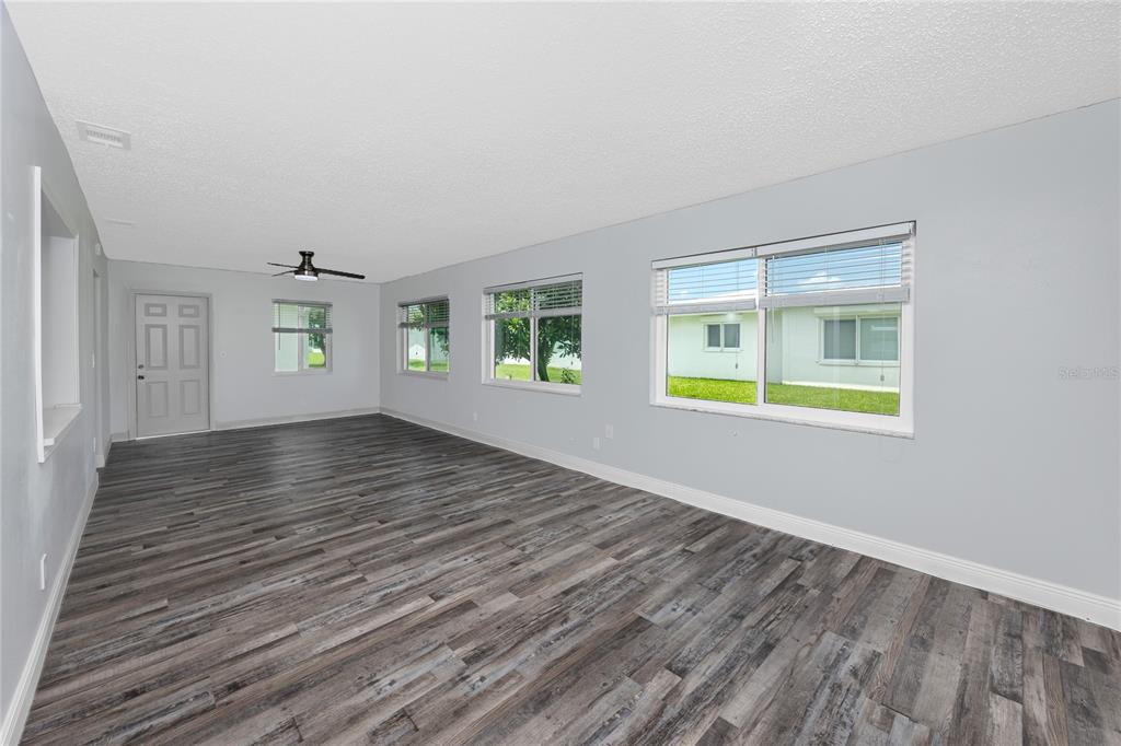 An empty room features gray walls, wood-look flooring, a ceiling fan, and multiple windows — 3455 100TH AVENUE N, PINELLAS PARK, FL