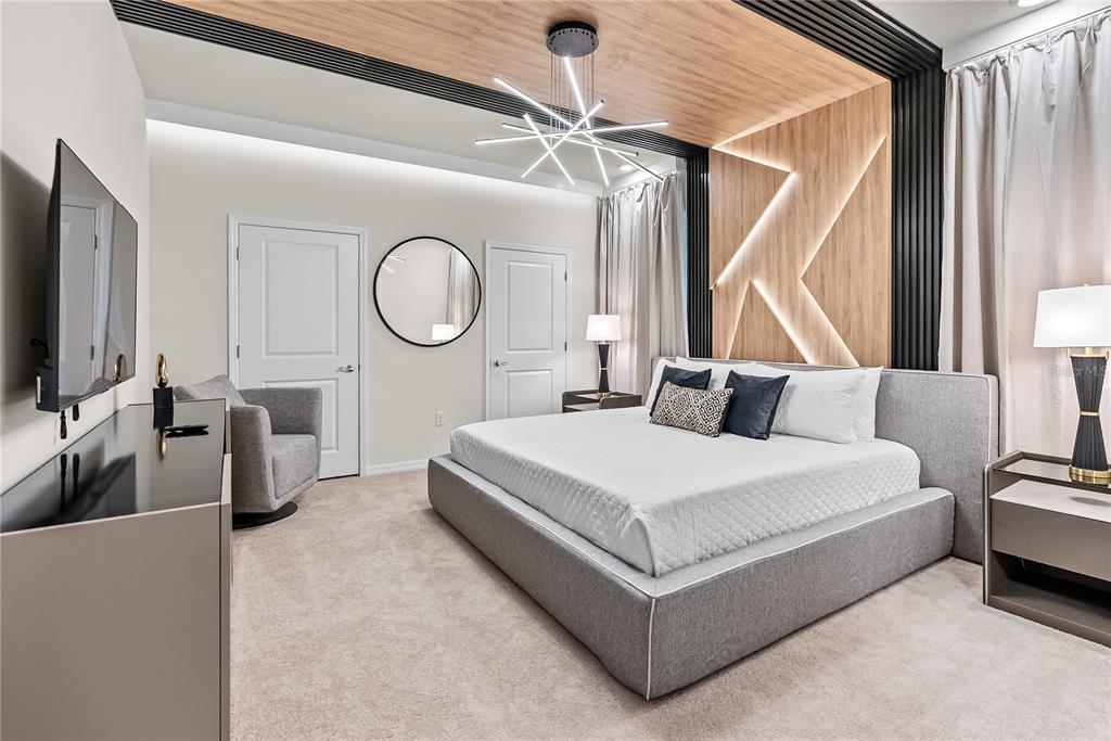 A modern bedroom features a gray upholstered bed, wood-paneled accent wall, and a geometric chandelier — 15969 CAPE CORAL LOOP, CLERMONT, FL