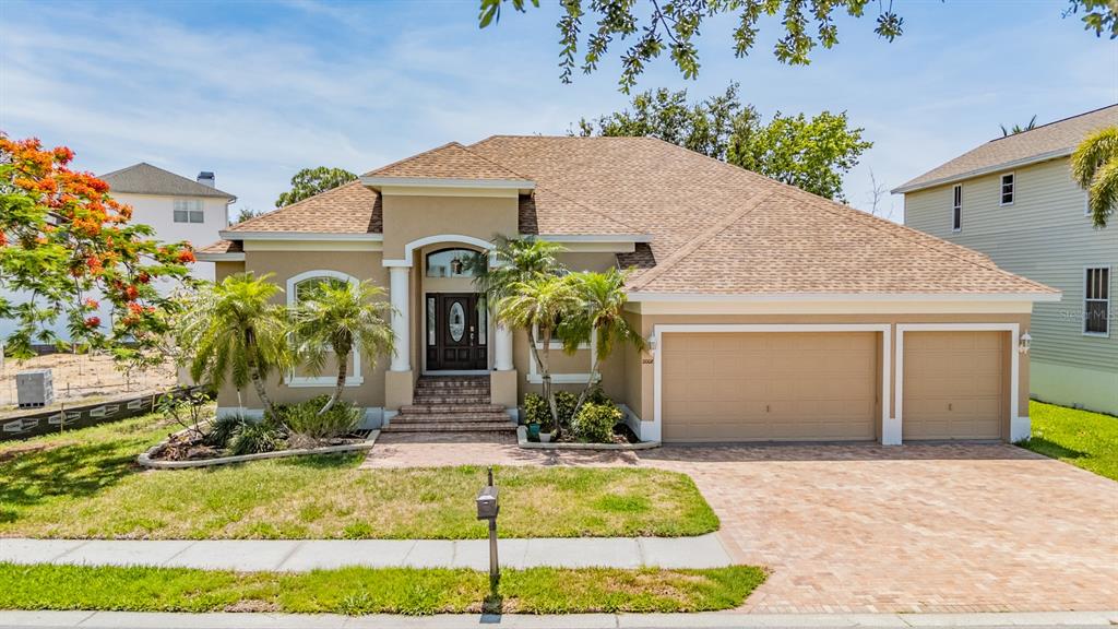 A tan single-story house with a brick driveway, three-car garage, and a large flowering tree — 2008 N POINTE ALEXIS DRIVE, TARPON SPRINGS, FL