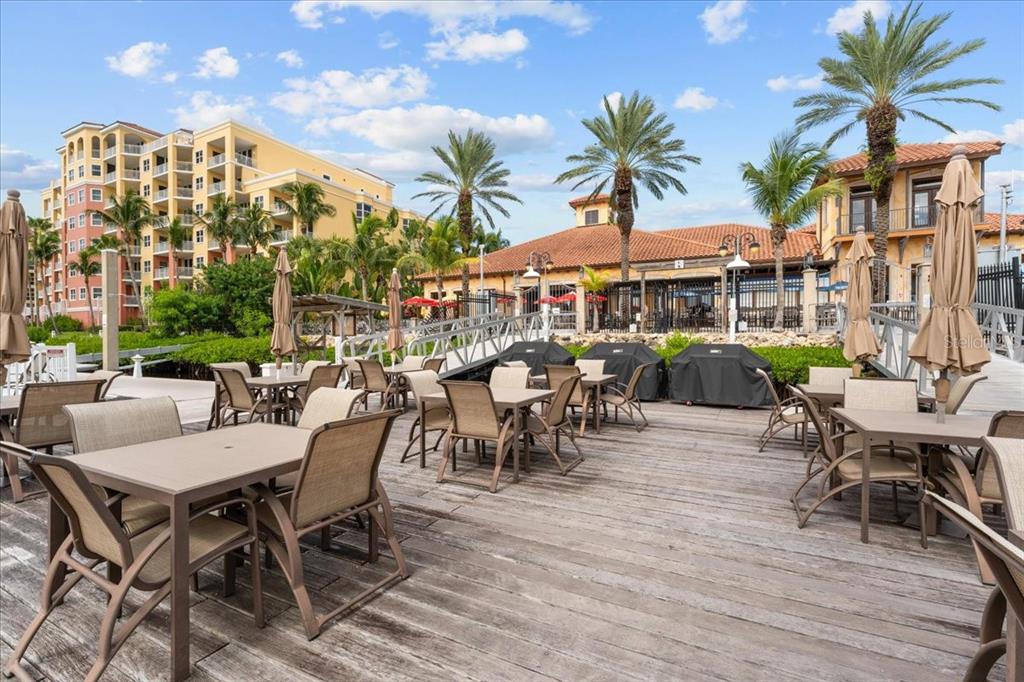 An outdoor wooden deck features dining tables and chairs near a building and palm trees — 102 RIVIERA DUNES WAY UNIT C-12, PALMETTO, FL