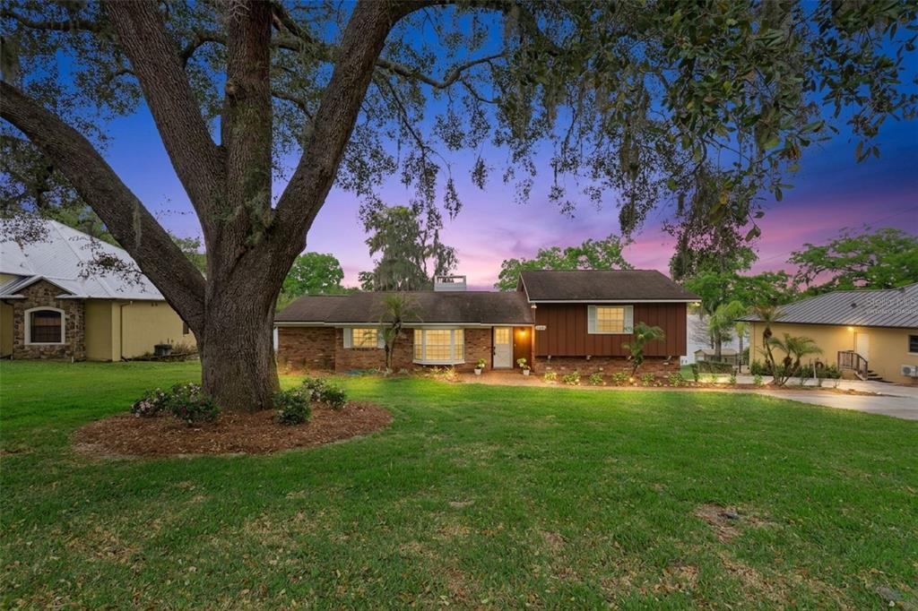 A single-story brick and wood-sided house sits behind a large oak tree at dusk — 2874 E CROOKED LAKE DRIVE, EUSTIS, FL