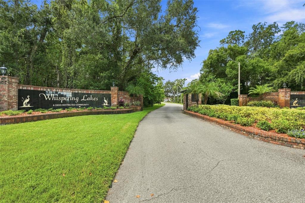 A brick entrance sign for Whispering Lakes community stands beside a paved road and trees — 501 MANISHA PLACE, TARPON SPRINGS, FL