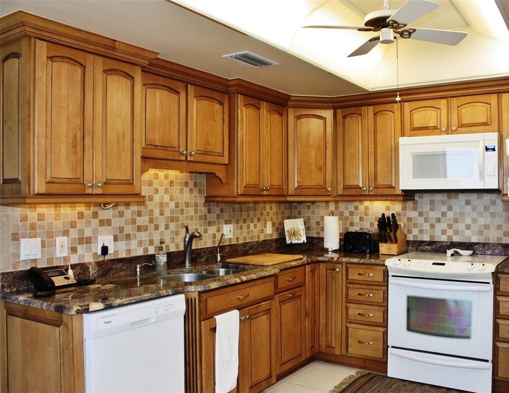 A kitchen features wood cabinets, granite countertops, a tiled backsplash, and white appliances — 150 BELLEVIEW BOULEVARD UNIT 507, BELLEAIR, FL