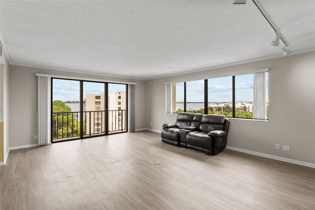 A living room with light wood-look flooring, a dark leather sofa, and large windows overlooking water — 150 BELLEVIEW BOULEVARD UNIT 507, BELLEAIR, FL