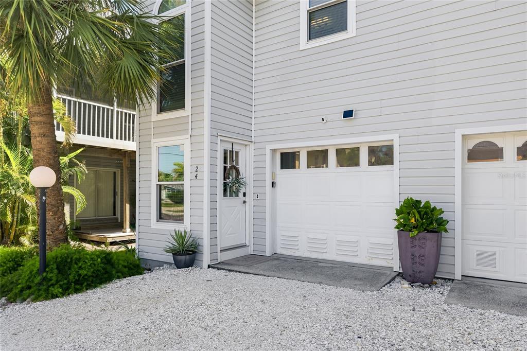 A two-story house exterior features light gray siding, a white garage door, and a gravel driveway — 6250 HOLMES BOULEVARD UNIT 24, HOLMES BEACH, FL