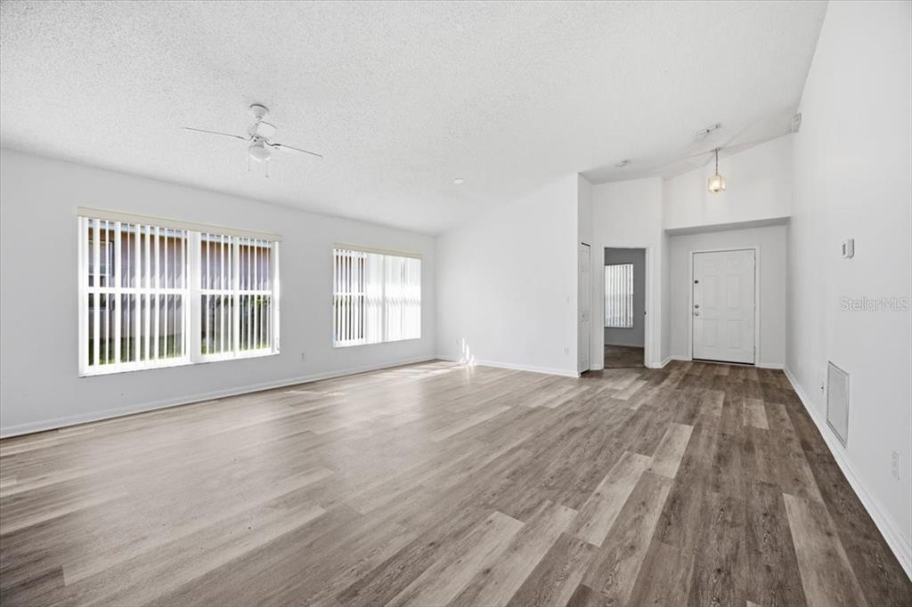 An empty living room features white walls, wood-look flooring, a ceiling fan, and large windows — 15702 BAY VISTA DRIVE, CLERMONT, FL