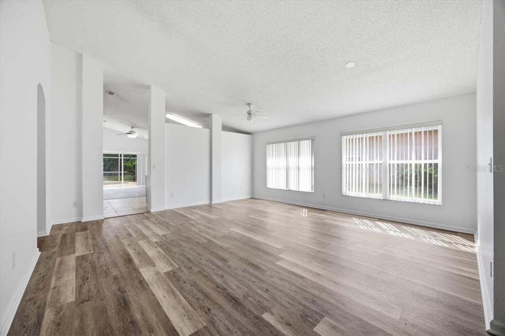 An empty living room with white walls, wood-look flooring, and large windows with vertical blinds — 15702 BAY VISTA DRIVE, CLERMONT, FL