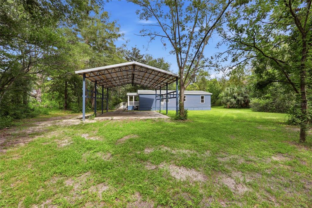 A blue mobile home sits behind a large metal carport on a grassy, wooded lot — 7412 NW STATE ROAD 45, HIGH SPRINGS, FL