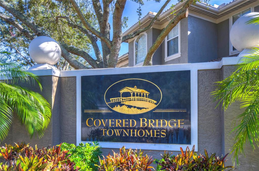 A sign for Covered Bridge Townhomes stands in front of a building with palm trees — 3292 COVERED BRIDGE DRIVE E, DUNEDIN, FL