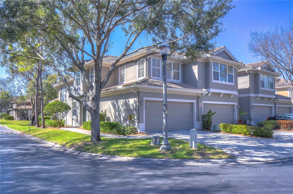 A two-story townhouse with a tan exterior, two garage doors, and a large shade tree — 3292 COVERED BRIDGE DRIVE E, DUNEDIN, FL