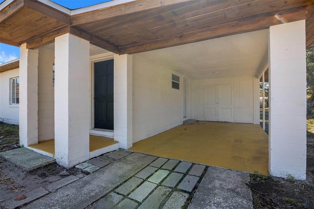 A covered carport with a yellow floor, white block pillars, and a dark front door — 6146 DAHLIA DRIVE, ORLANDO, FL