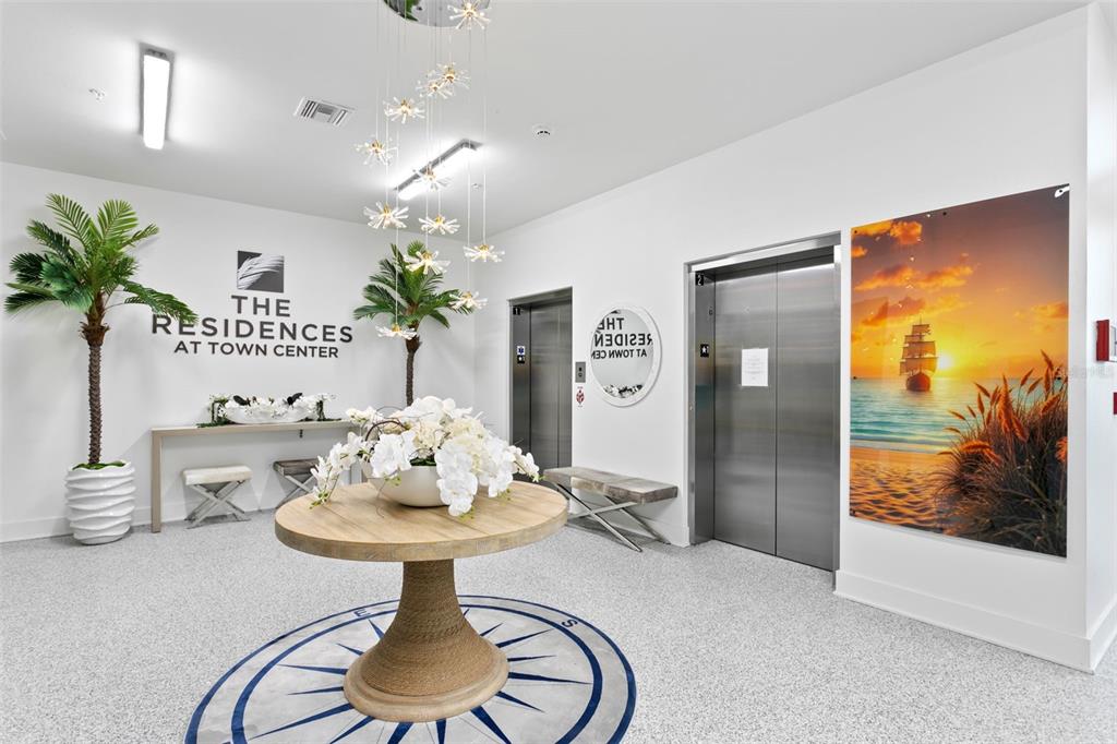 A bright apartment building lobby features a round table, two elevators, and a nautical-themed mural — 400 150TH AVENUE UNIT 205, MADEIRA BEACH, FL