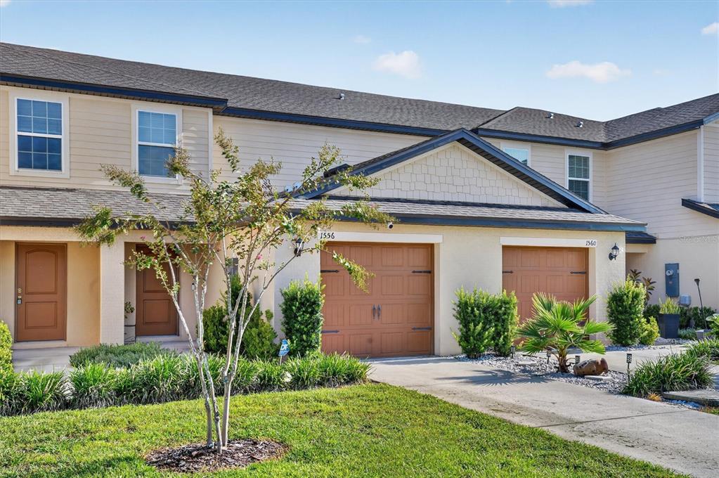 A two-story townhouse exterior features tan siding, brown garage doors, and a small front yard — 1556 WYNFORD CIRCLE, TAVARES, FL