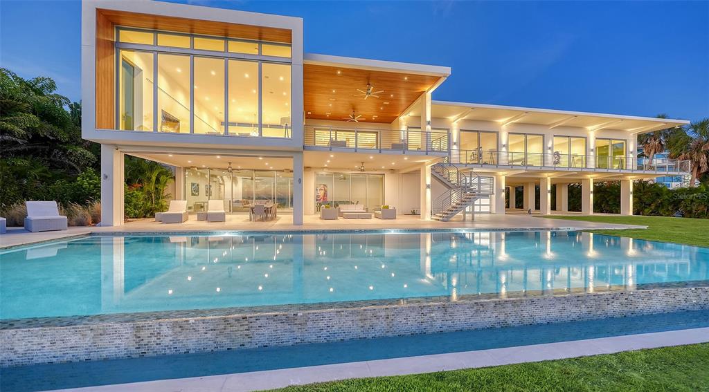 A modern two-story house with a large infinity pool and expansive glass walls at dusk — 4123 BAY SHORE ROAD, SARASOTA, FL