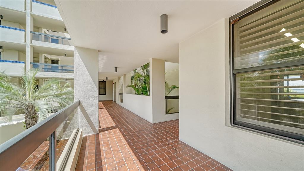 A tiled outdoor balcony walkway features a glass railing, white walls, and a large window — 1930 HARBOURSIDE DRIVE UNIT 122, LONGBOAT KEY, FL