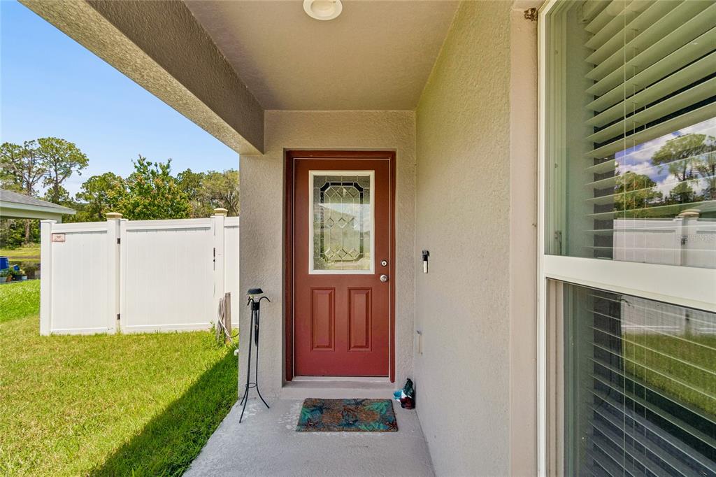A red front door with a glass insert sits under a covered porch beside a window — 6917 GIDEON CIRCLE, ZEPHYRHILLS, FL