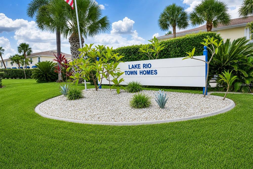 A white sign reading Lake Rio Town Homes sits in a landscaped bed with gravel — 26310 RAMPART BOULEVARD UNIT D504, PUNTA GORDA, FL