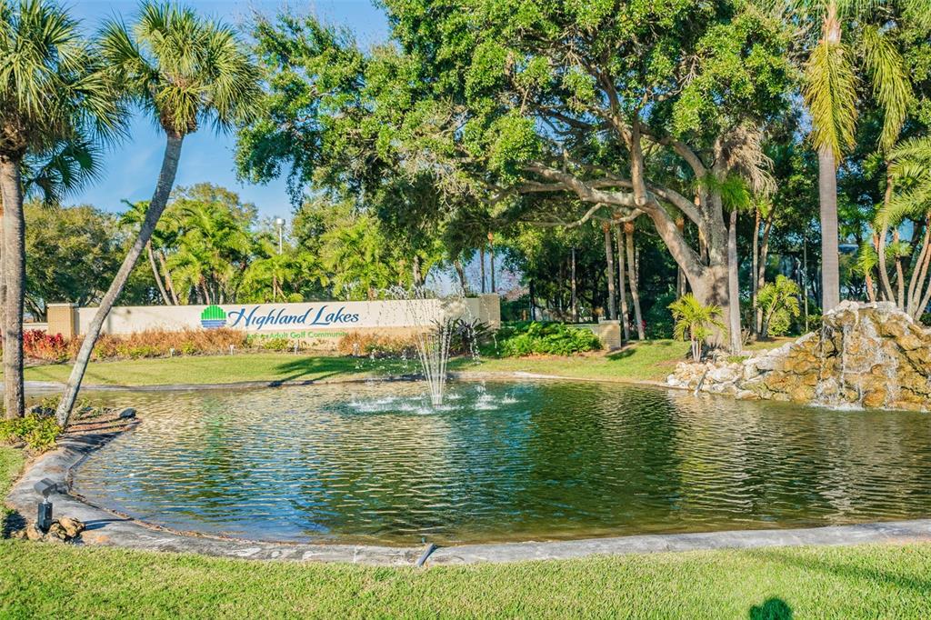 A pond with a fountain sits in front of a Highland Lakes community entrance sign — 2150 SHELLY DRIVE UNIT B, PALM HARBOR, FL
