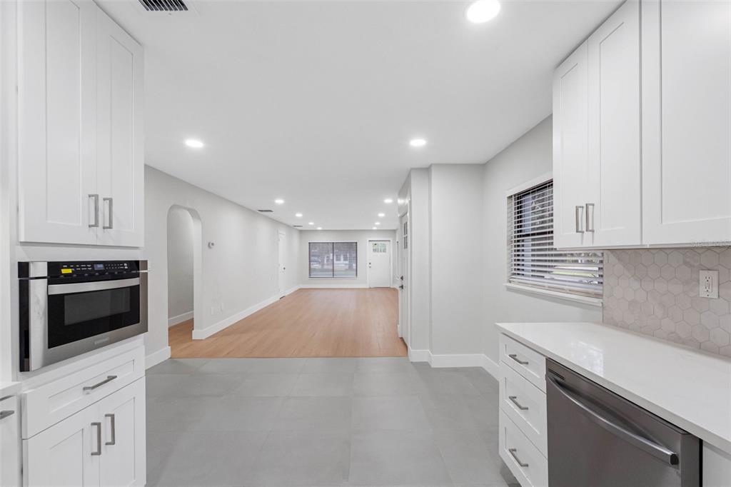 A kitchen with white cabinets and gray tile flooring opens into a light-filled living area — 1005 APACHE TRAIL, CLEARWATER, FL