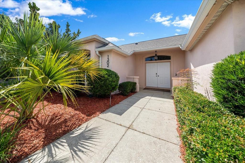A concrete walkway leads to the double front doors of a light-colored stucco house — 1302 LOPEZ LANE, THE VILLAGES, FL