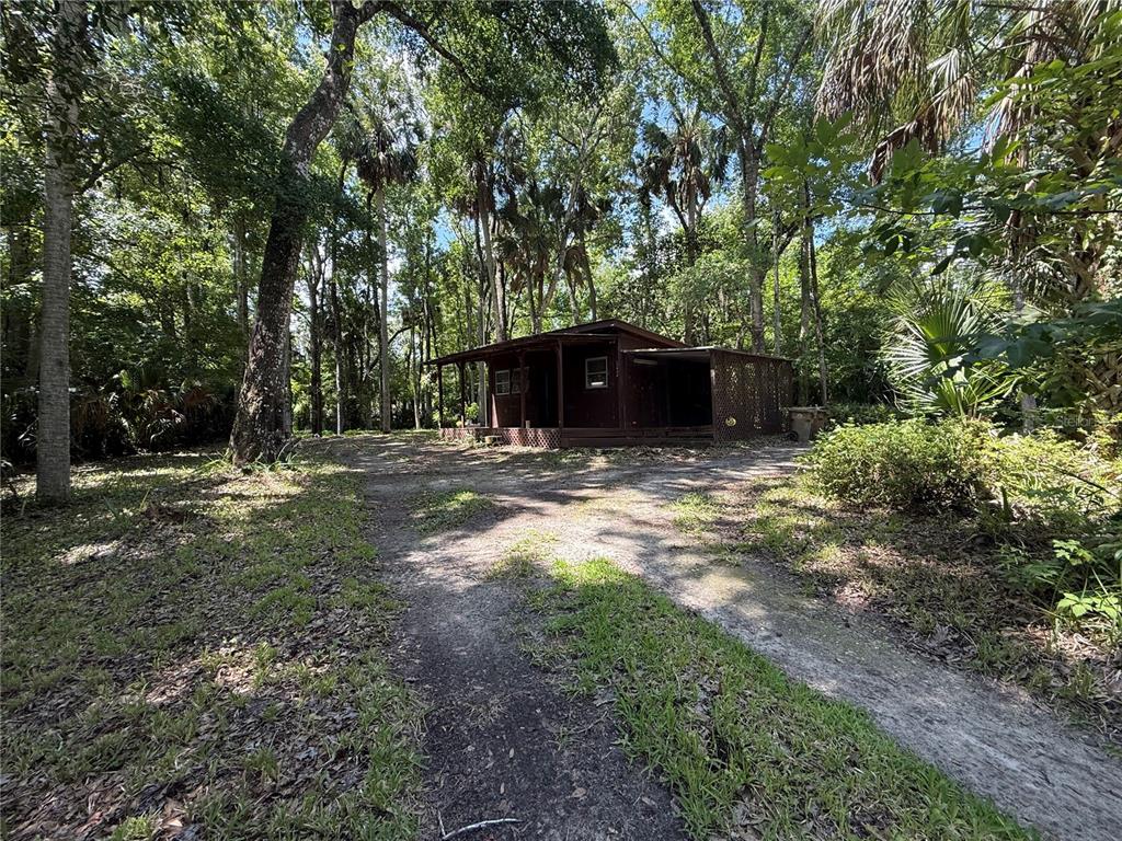 A small, dark-brown wooden cabin sits in a wooded area with a dirt driveway — 23946 INDIGO ROAD, ASTOR, FL