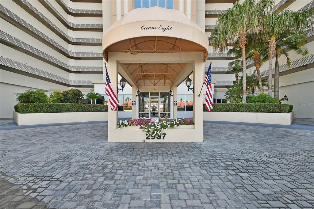 The exterior entrance of a multi-story building features a covered driveway, flags, and landscaping — 2937 S ATLANTIC AVENUE UNIT 708, DAYTONA BEACH SHORES, FL