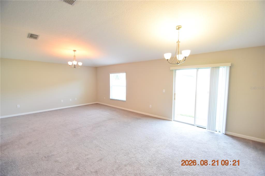 An empty living room features beige walls, neutral carpeting, two chandeliers, and a sliding glass door — 8436 HUNTERS FORK LOOP, RUSKIN, FL