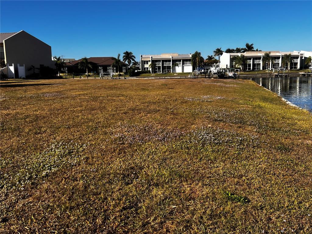 A vacant grassy lot sits alongside a canal with residential buildings in the background — 1443 MEDITERRANEAN DRIVE, PUNTA GORDA, FL