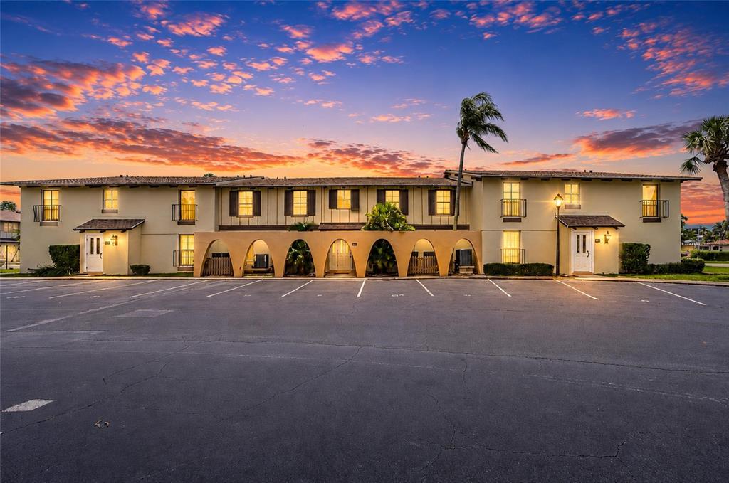 A two-story apartment building with arched ground-level walkways stands before a parking lot at sunset — 501 PLAZA SEVILLE COURT UNIT 2, TREASURE ISLAND, FL