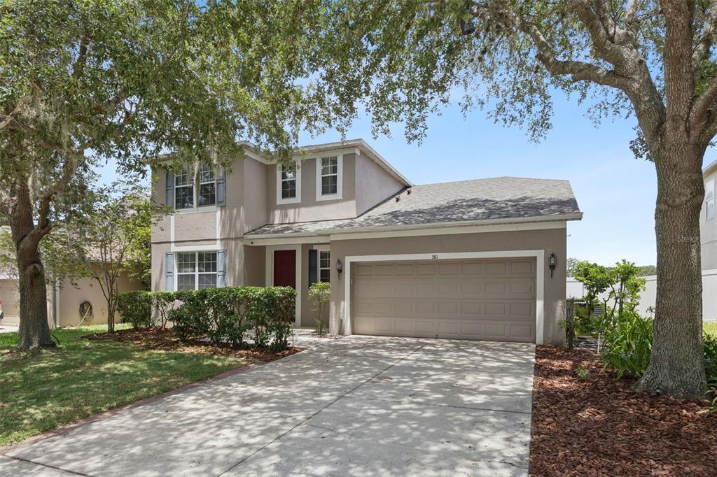 A two-story house with a beige stucco exterior, a two-car garage, and mature trees — 381 MISTY OAKS LANE, EUSTIS, FL
