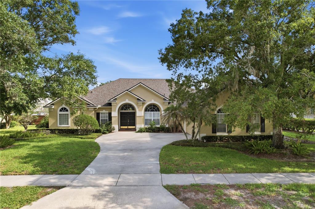 A single-story tan house with a concrete driveway and arched windows under large oak trees — 11457 SWIFT WATER CIRCLE, ORLANDO, FL