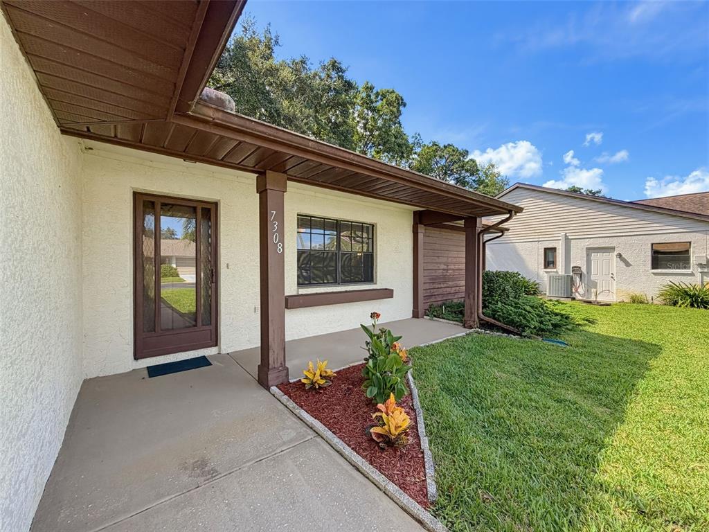 The exterior of a house features a covered porch, a front door, and landscaped garden beds — 7308 BALTUSROL DRIVE, NEW PORT RICHEY, FL