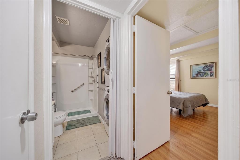 A bathroom with a shower and stacked laundry units sits beside a bedroom with wood flooring — 1513 CLUB CIRCLE UNIT 1513, LAKESHORE, FL